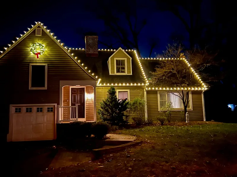 Cape Cod home at night with warm white lights along every roofline and dormer and a lit wreath on the gable