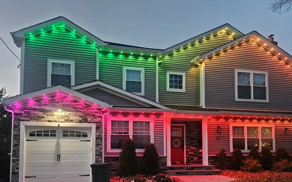 Two-story colonial at dusk with permanent roofline lights shading from green on the upper roof to pink and red over the porch