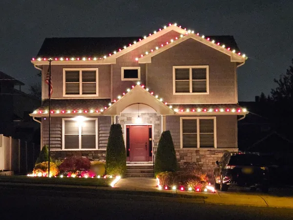 Two-story home at night with alternating red and white bulbs along the rooflines and entry gable and lights edging the garden beds